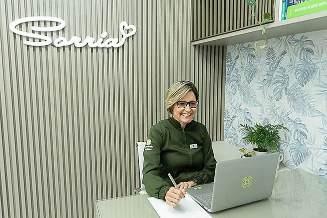 Paper free. Dr.a Claudia Assis sentada na sua mesa consultando o sistema Clinicorp,
