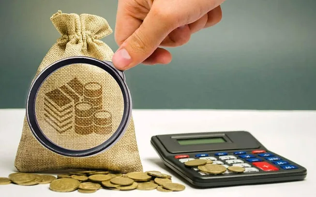 Elisão fiscal odontologia: imagem ilustrativa de uma pessoa com uma lupa, observando um saco de dinheiro posto ao lado de uma calculadora, com algumas moedas espalhadas pela mesa.