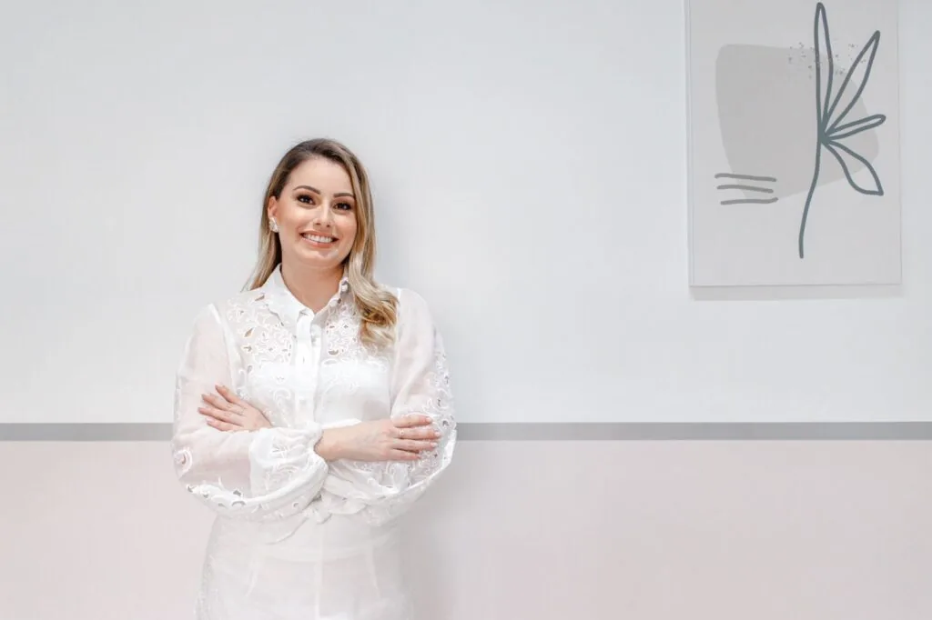Dentista de jaleco branco, de braços cruzados, em frente a uma parede clara