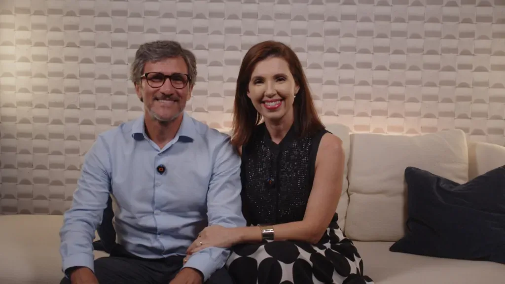 Imagem da Dra. Bárbara e Dr. Edival, da clínica Nouva New Smile. Ambos estão sentados no sofá da clínica e sorriem para a câmera.
